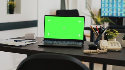 Office with green screen laptop on desk and monitor in background with stock exchange listings. Mockup notebook in workspace and computer screen in background displaying trading platform
