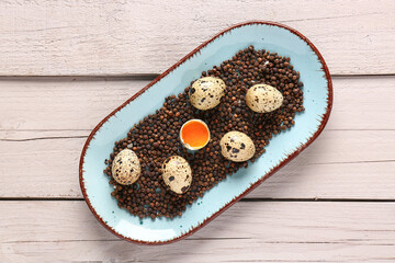 Plate with black peppercorns and raw quail eggs on white wooden background