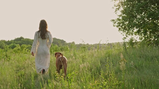 young cheerful woman playing with her dog outdoors in beautiful countryside field. Female and ridgeback dog walking and having fun