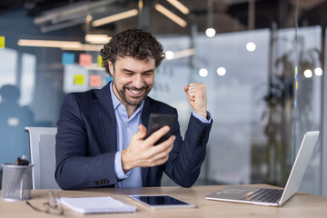 Happy businessman in suit celebrating success on smartphone in modern office. Professional expressing excitement and joy at desk with laptop, notepad, and glasses. Concept of business success