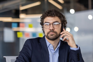 Businessman in office talking on smartphone, wearing glasses and suit. Confident professional working in modern office environment with colorful sticky notes in background. Concept of communication