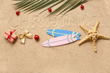 Starfish, toy surfboard, and palm branch with Christmas decor and word CHRISTMAS on sand. Top view