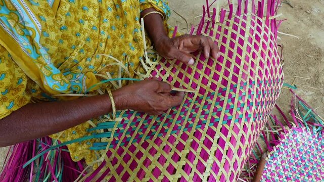 An old woman is making a colorful hand fan out of palm leaf fibers. Handicrafts are made by weaving colorful dry leaves.