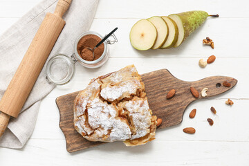 Wooden board with half of tasty pear strudel, cut fresh fruit and rolling pin on white wooden background
