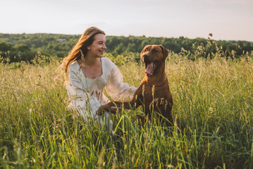 young cheerful woman playing with her dog outdoors in beautiful countryside field. Female and ridgeback dog walking and having fun