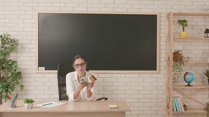 Woman college teacher sitting at desk in classroom in front of chalkboard, typing credit card...