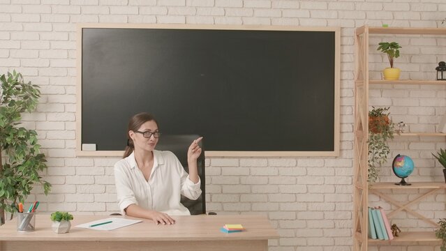 Woman college teacher sitting at desk in classroom in front of chalkboard pointing at empty area and smiling. Education concept.