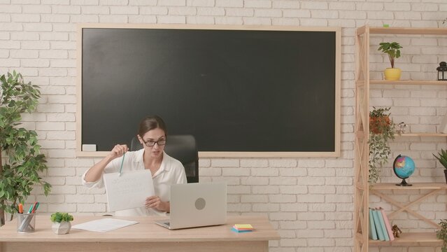 Woman college teacher sitting at desk in classroom in front of chalkboard explaining lesson for students on laptop online. Education concept.