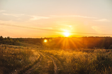 a dirt road turning left into a field, against the backdrop of a bright sunset with light orange rays