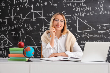 Mature female math teacher sitting at table with laptop and globe near blackboard in classroom