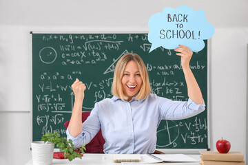 Happy mature female math teacher holding paper sheet with text BACK TO SCHOOL in classroom