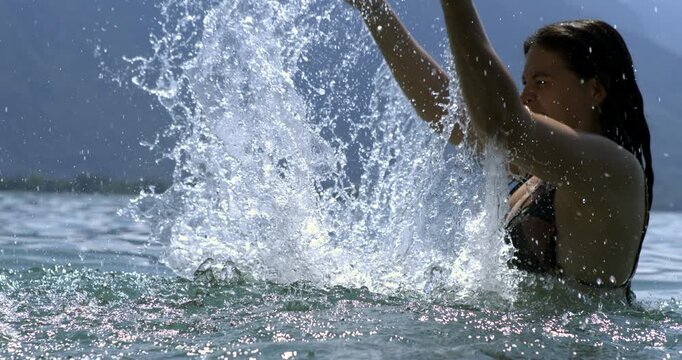 Woman raising arms and splashing water in slow-motion at 800 fps, showcasing the vibrant motion and fun in a serene lake environment