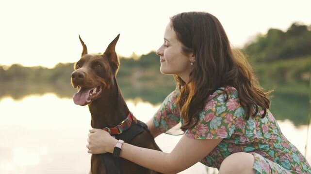 young woman playing with her cute doberman dog outdoors near the lake during sunset