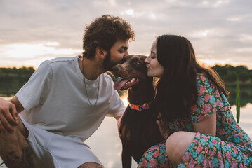 young happy couple having picnic and playing with their doberman dog outdoors near the lake during beautiful sunset