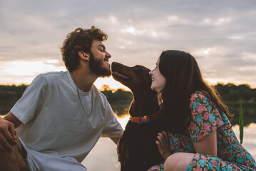 young happy couple having picnic and playing with their doberman dog outdoors near the lake during beautiful sunset