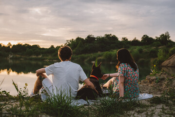 young happy couple having picnic and playing with their doberman dog outdoors near the lake during beautiful sunset