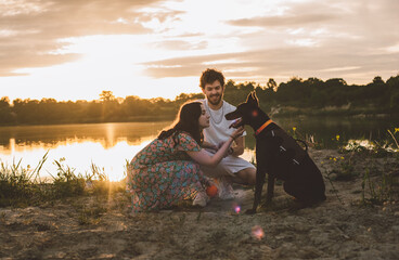 young happy couple having picnic and playing with their doberman dog outdoors near the lake during beautiful sunset