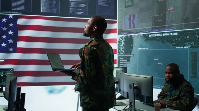 Professionals in US military uniforms operate computers and big screen, working on surveillance and intelligence to ensure national security from a high tech control center. Camera B.