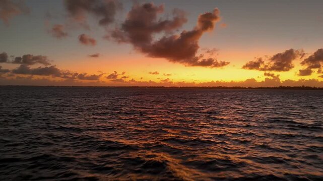 Drone flying over the ocean water under vibrant colorful orange sunrise sky and clouds near the tropical island of Bora Bora in French Polynesia. 