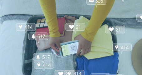 Image of media notifications over biracial woman with smartphone packing suitcase