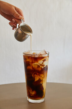 Pouring milk over a glass of iced coffee on top pf a concrete table.