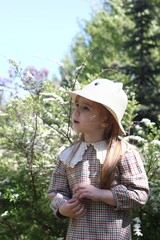 Adorable little girl in a lovely pink and white dress and hat standing in a vibrant flower garden.