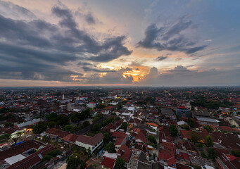 The photo captures an urban landscape during sunrise or sunset. The dramatic sky frames densely packed buildings, their roofs varying in shades of red and brown