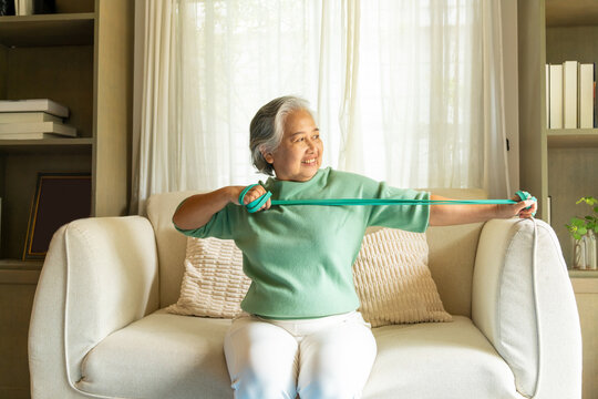 Happy Asian senior mature woman enjoy healthy lifestyle resistance band training exercise for stretching body muscle shoulder and arm on sofa in living room. Elderly self-care and home fitness concept