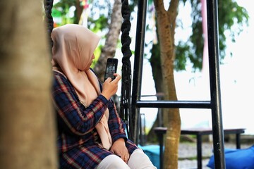 A Muslim woman is sitting in nature relaxing holding her cell phone