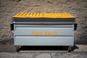 The words Food Waste stenciled in yellow on a large commercial trash bin near a restaurant.  The...