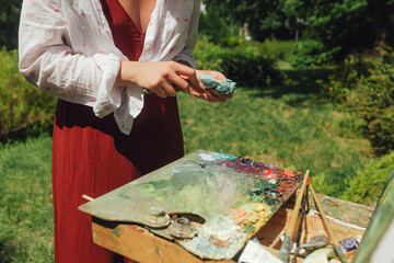 Close up of woman's hands with brushes and paints near a canvas with etude. Mindfulness, art therapy, creativity concept. Process of plein air painting in nature. Fine art, sketch, sketchbook
