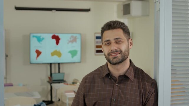 Portrait of young confident Caucasian secondary school male teacher standing in classrom, putting off glasses, smiling and posing for camera
