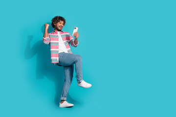 Photo of overjoyed glad young man wear stylish striped clothes hold modern gadget empty space isolated on cyan color background