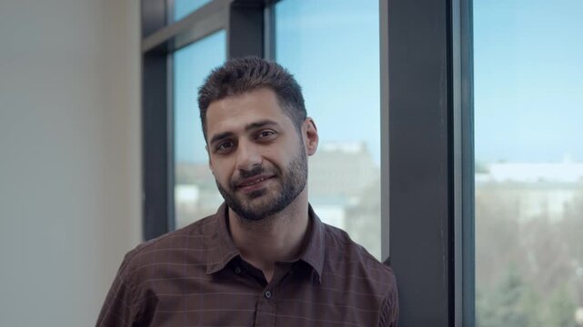 Portrait of young Caucasian male school principal with confident smile standing leaning against window looking directly at camera