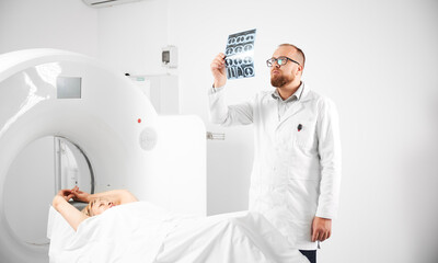 Obraz premium Medical computed tomography or MRI scanner. Doctor holding and examining results of MRI. Woman patient lying on CT scan bed. Concept of medicine, healthcare and modern diagnostics.