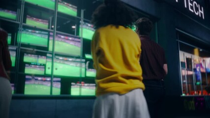 Young Group of Men and Women Enthusiastically Watches a Soccer Match on a Storefront TV Display Late in the Evening. Community of Sports Fans Excited to Support Their National Football Team. Back View