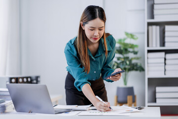 Asian woman sit at workplace desk holds cellphone staring at laptop, synchronize data between...