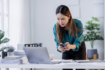 Asian woman sit at workplace desk holds cellphone staring at laptop, synchronize data between computer and gadget in office, use corporate devices and business application, plan work, use organizer