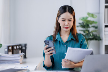 Asian woman sit at workplace desk holds cellphone staring at laptop, synchronize data between...