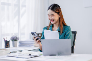 Asian woman sit at workplace desk holds cellphone staring at laptop, synchronize data between...