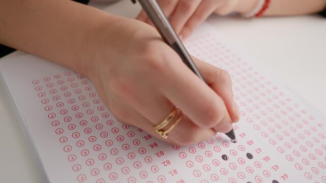 Female circles correct answers in standard exam test paper with black pen. Student filling up the answer sheet, coloring right answer, close up shot.