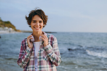 Young smiling happy woman wear shirt casual clothes hold headphones on neck listen to music look camera rest on sea ocean sand shore beach outdoor seaside in summer day free time. Lifestyle concept.