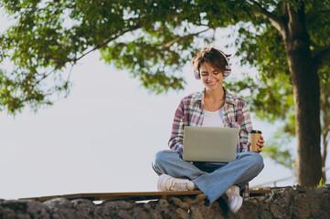 Full body young smiling woman wear shirt casual clothes work hold use laptop pc computer drink coffee sits walk rest relax in spring sunshine green city park outdoors on nature. Urban leisure concept.