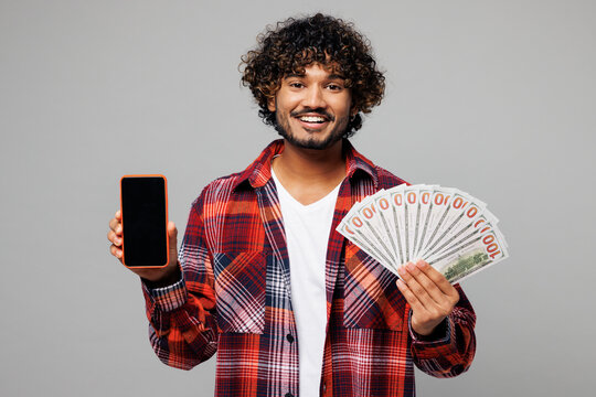 Young happy Indian man he wear red shirt casual clothes use blank screen mobile cell phone hold fan of cash money in dollar banknotes isolated on plain grey color background studio. Lifestyle concept