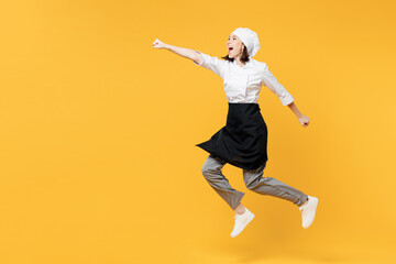 Fototapeta premium Full body young fun chef cook baker woman wear white shirt black apron uniform toque hat jump high do super hero gesture, clench fist isolated on plain yellow background studio. Cooking food concept.