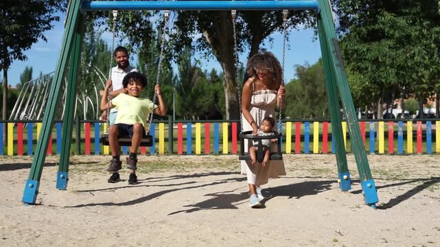 Latin family with afro hair enjoying a day at the playground. Father and mother pushing their son and baby on the swings