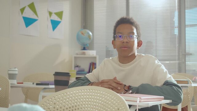 Portrait of serious teenage African American boy wearing glasses sitting at desk in classrom at school during recess time and looking at camera