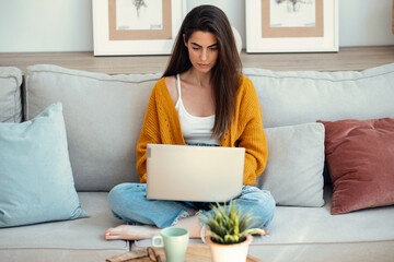 Beautiful young woman working with her laptop while sitting on a couch at home.