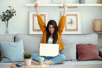 Pretty motivated woman celebrating something while working with laptop sitting on a couch at home