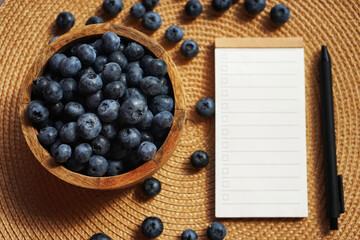 Fresh blueberries in a wooden bowl next to a notepad with a list on a beige napkin